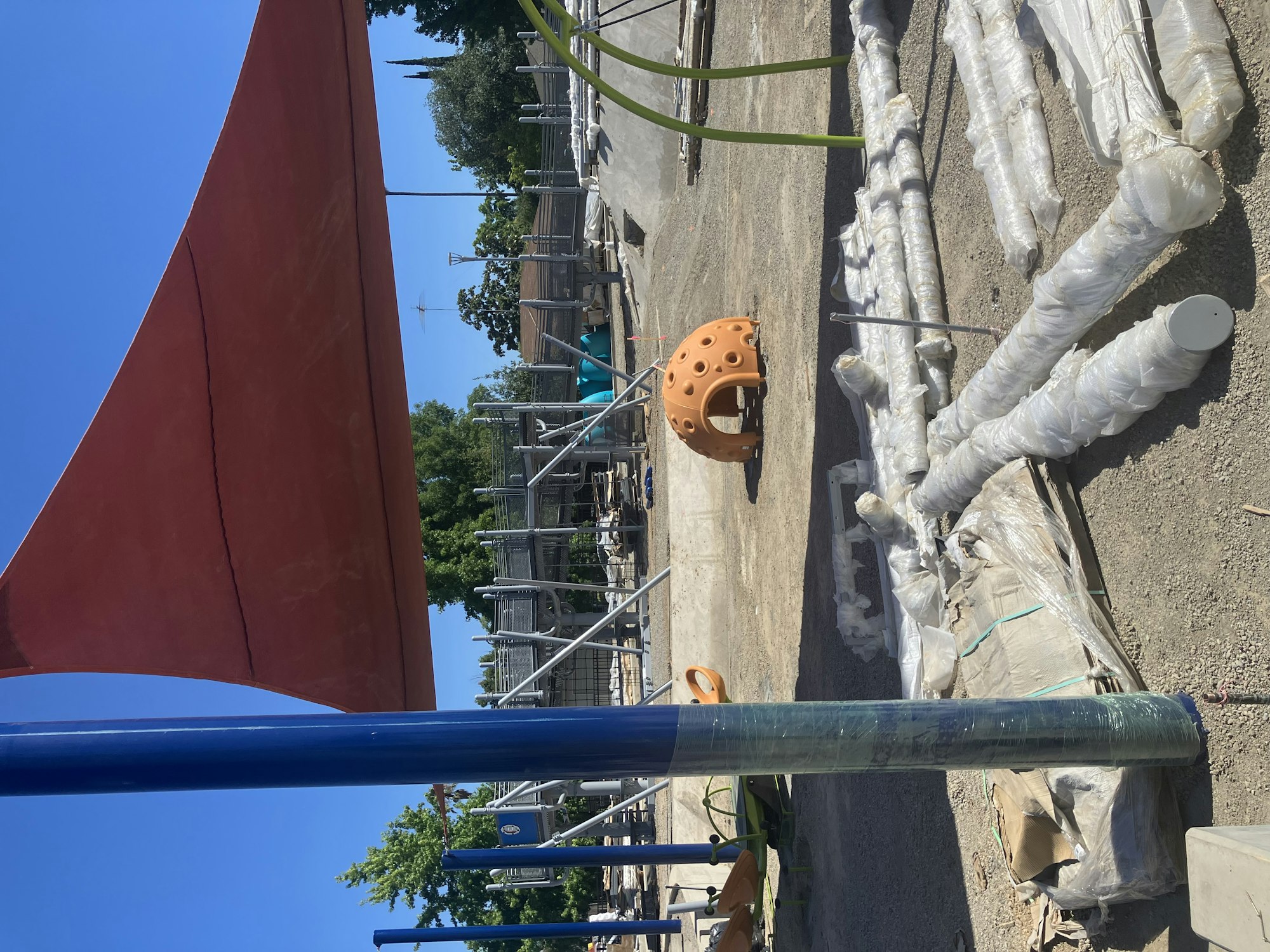 The image shows a playground area with construction materials, including pipes and an orange climbing structure under a shade.
