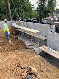 A person in a safety vest and helmet is standing at a construction site with scaffolding and cinder block walls.