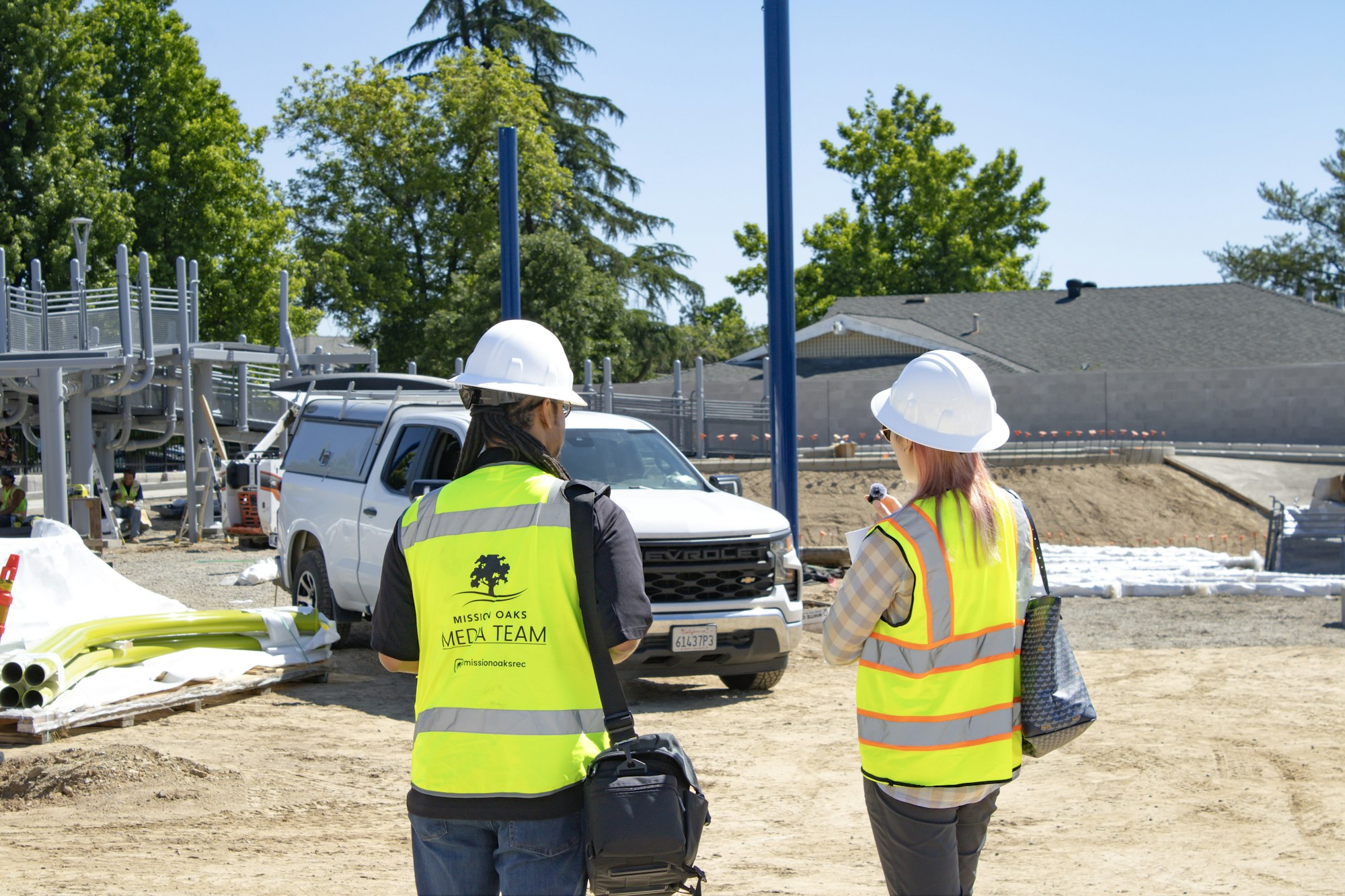 Two people in safety gear are discussing at a construction site, with construction vehicles and materials visible in the background.
