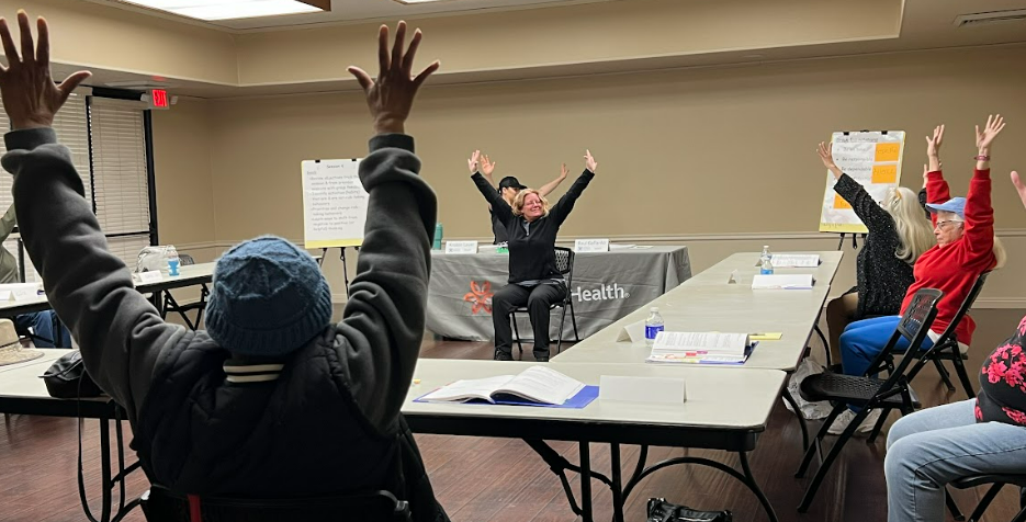 People with raised hands in a meeting or workshop, sitting at tables with papers and a flip chart in the background.