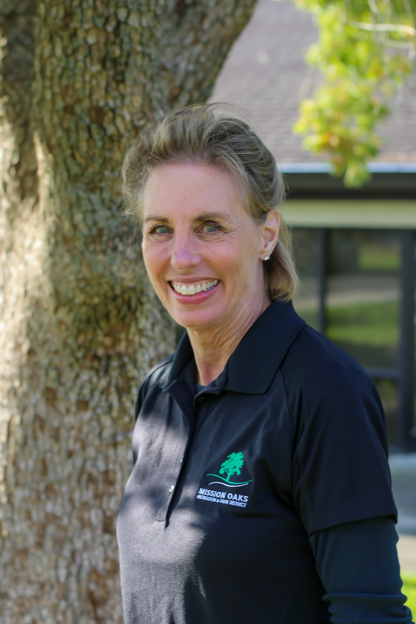 A person wearing a black "Mission Oaks" polo shirt, smiling outdoors with a tree in the background.