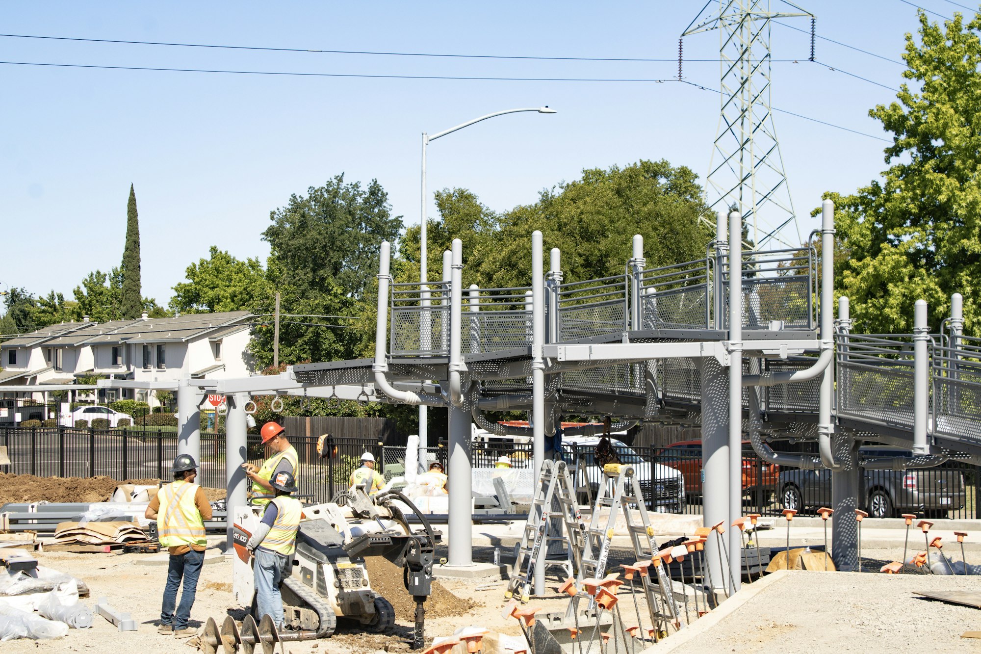 Construction workers are building a pedestrian bridge, with equipment and materials visible on-site. It’s a sunny day.