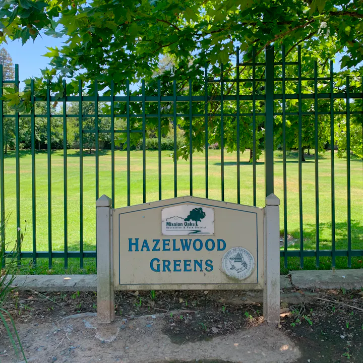 A sign marking "Hazelwood Greens," part of Mission Oaks Recreation & Park District, surrounded by green grass and trees.