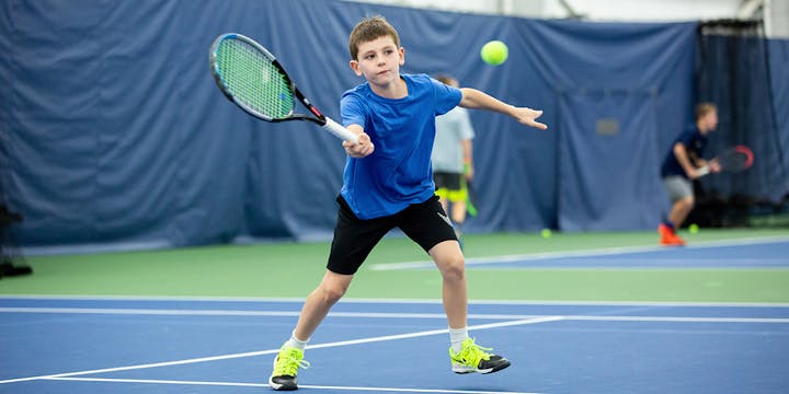 A child in a blue shirt playing tennis indoors, hitting a ball with a racquet.