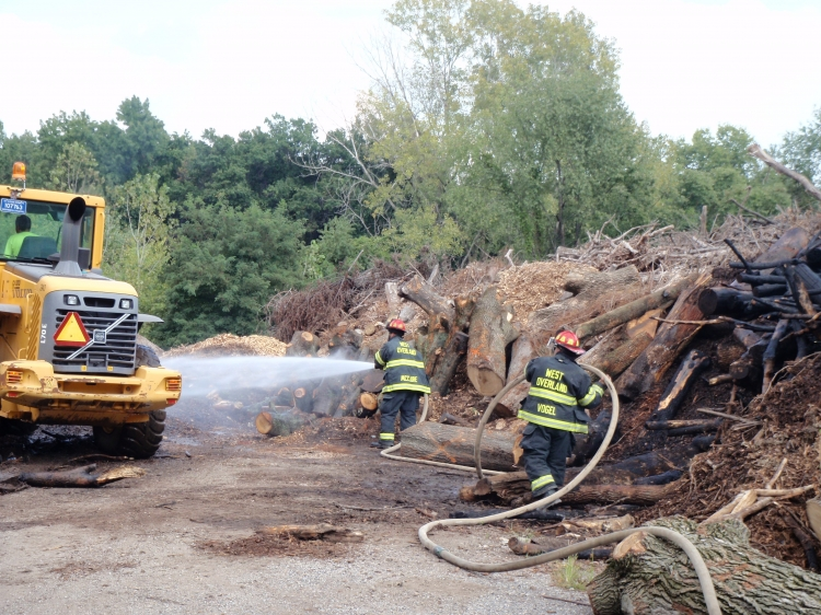 Firefighters spraying water on a pile of logs near a bulldozer in a wooded area.