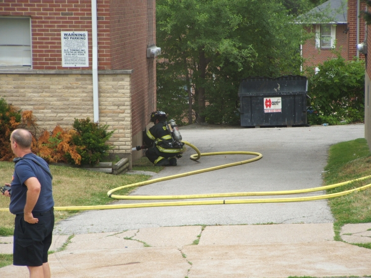A firefighter crouches with a hose near a building, while another person observes. A "No Parking" sign and dumpster are visible.