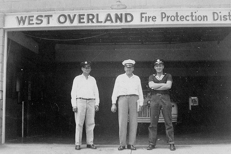 Three men stand in front of a fire station labeled "West Overland Fire Protection District" with a vehicle partially visible behind them.