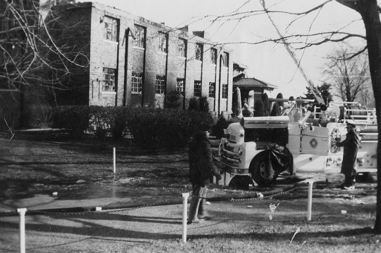 A vintage fire truck parked in front of a brick building, with firefighters and onlookers nearby.