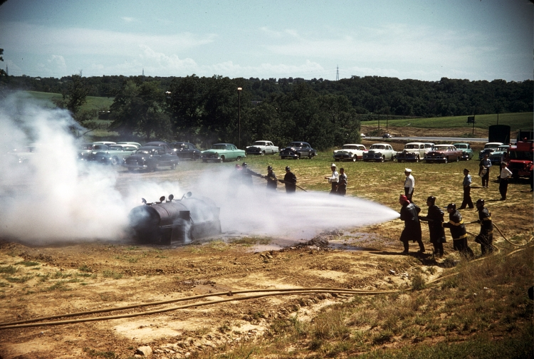 Firefighters spraying water on a burning object with cars in the background.