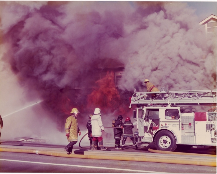 Firefighters battling a large blaze with hoses and a fire truck, amidst heavy smoke and flames.