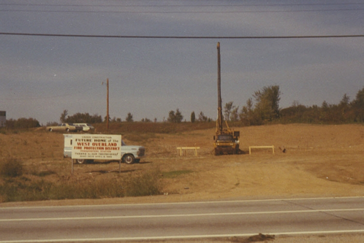 Construction site with a sign, truck, and drilling machine on a dirt field, bordered by a road and trees in the background.