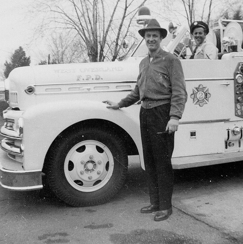 Two men with a vintage fire truck; one standing and one in the driver's seat, labeled "West Overland F.P.D."