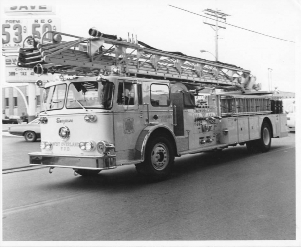 A vintage fire truck with a ladder is parked on a street near a gas station displaying prices.