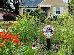 A garden with various flowers and a "Bee City" sign, in front of a small house with a gray roof and white window shutters.