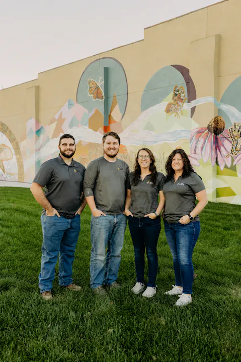 A group of four individuals stands in front of a colorful mural on a grassy area, all wearing matching gray shirts.