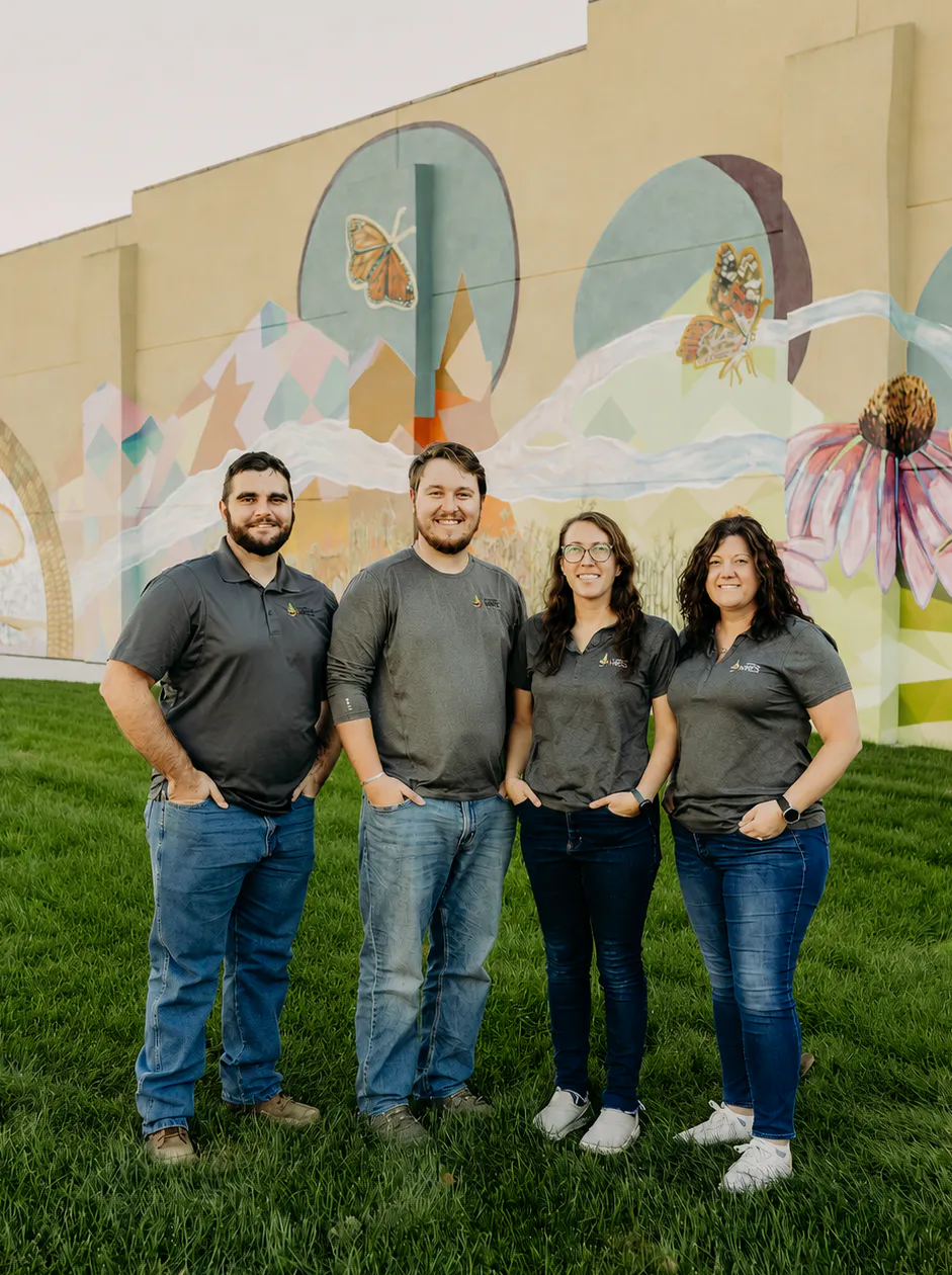 A group of four individuals stands in front of a colorful mural on a grassy area, all wearing matching gray shirts.