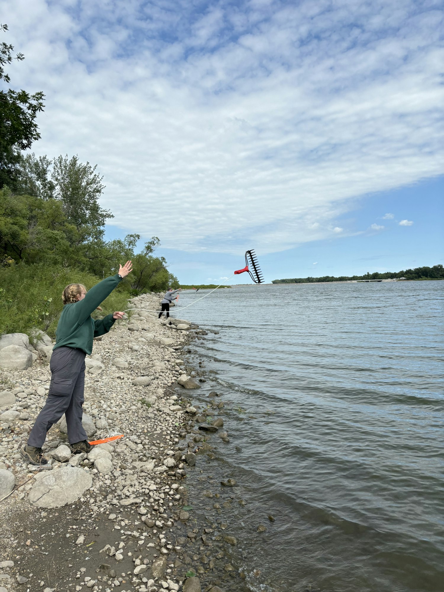 A person on a rocky lakeshore is flying a rake-shaped kite into the sky.