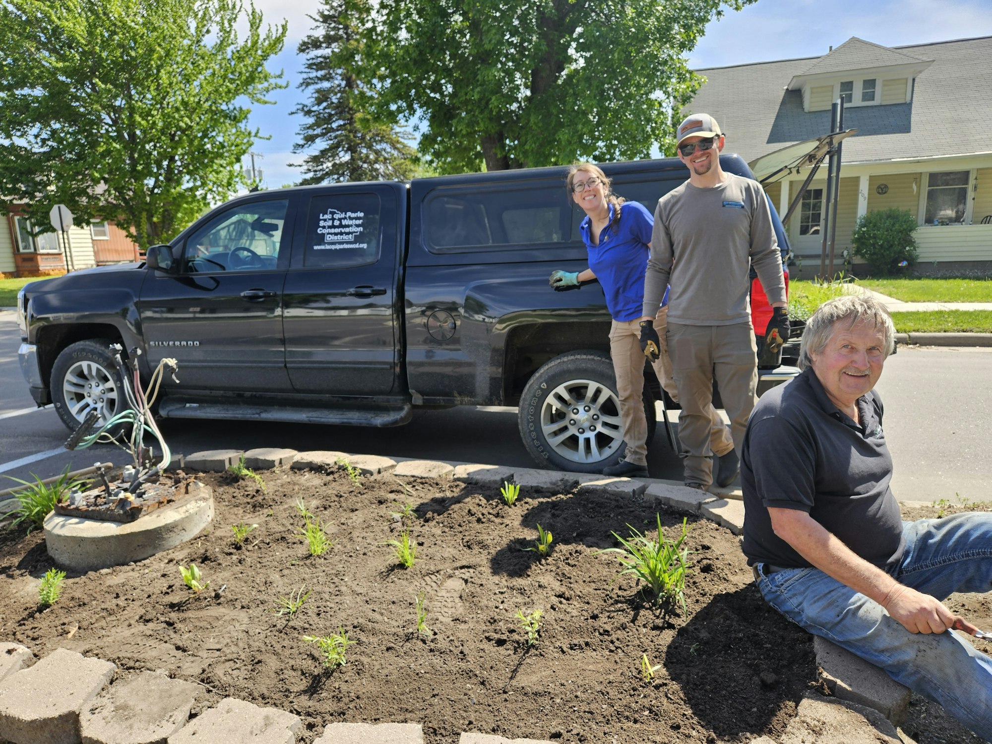 Three people planting in a garden bed near a black truck on a sunny day.