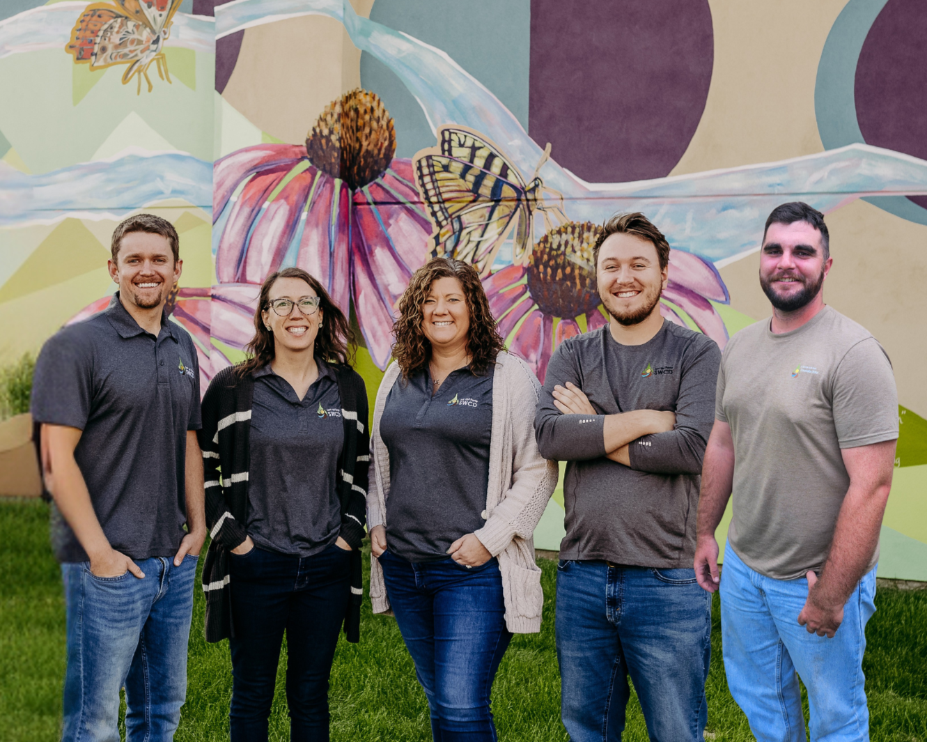 Five people smiling in front of a colorful mural with flowers and butterflies.