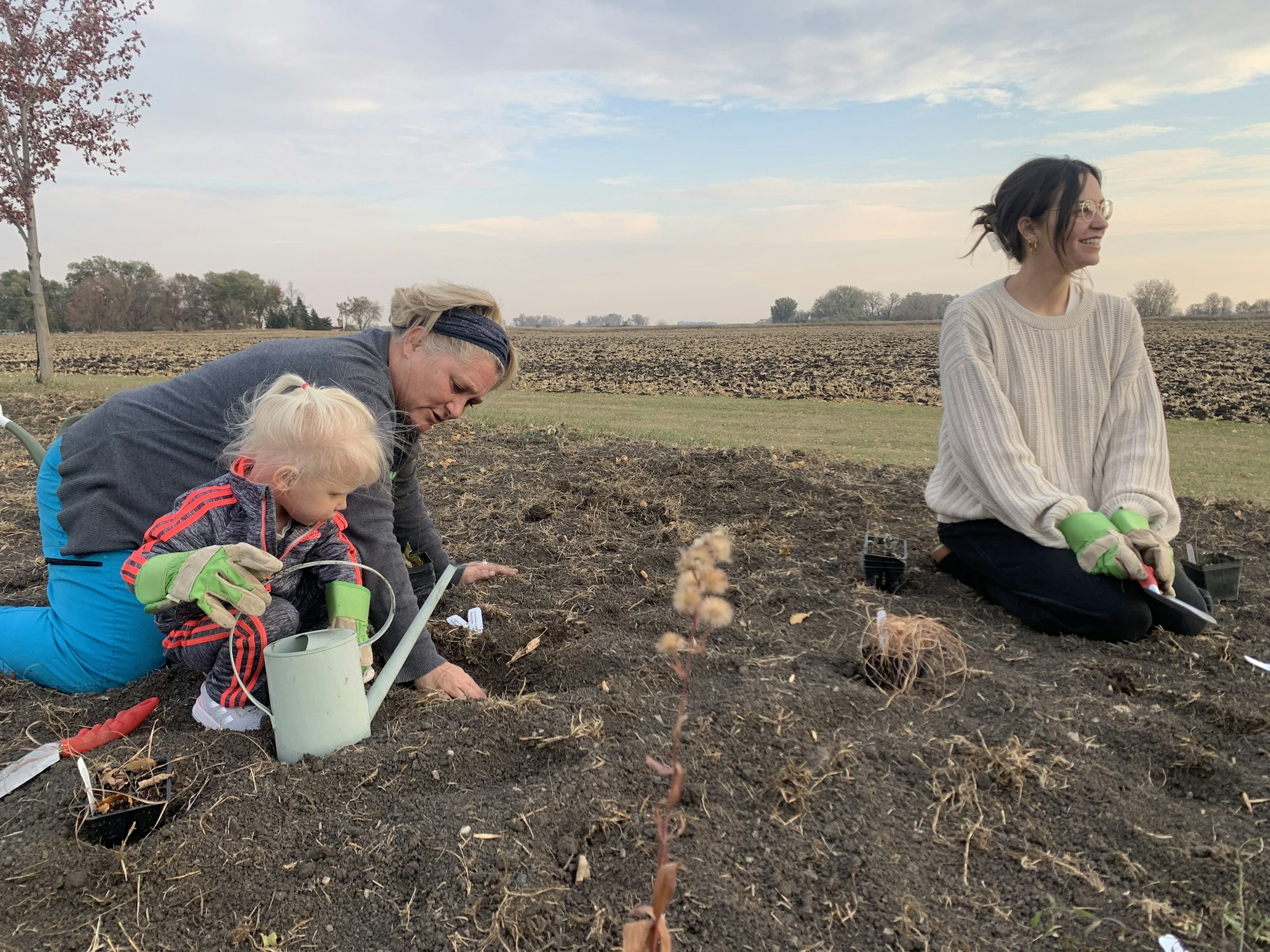 Three people gardening in a field; one woman planting with a child, another sitting nearby.