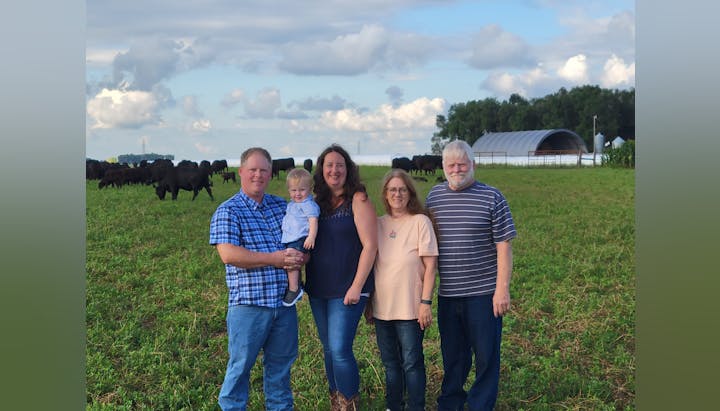Five people posing in a field with cows in the background under a cloudy sky.
