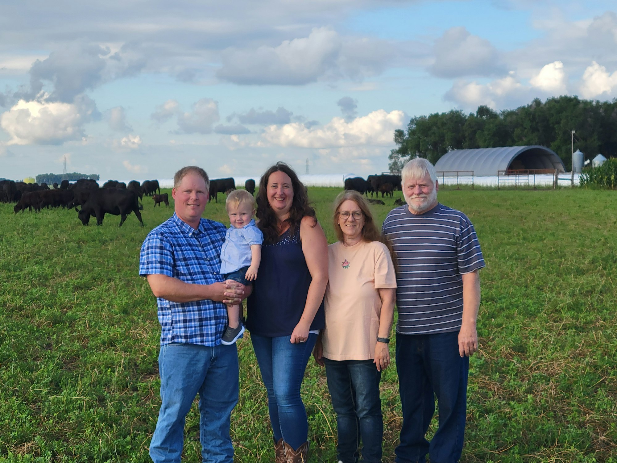 Five people posing in a field with cows in the background under a cloudy sky.