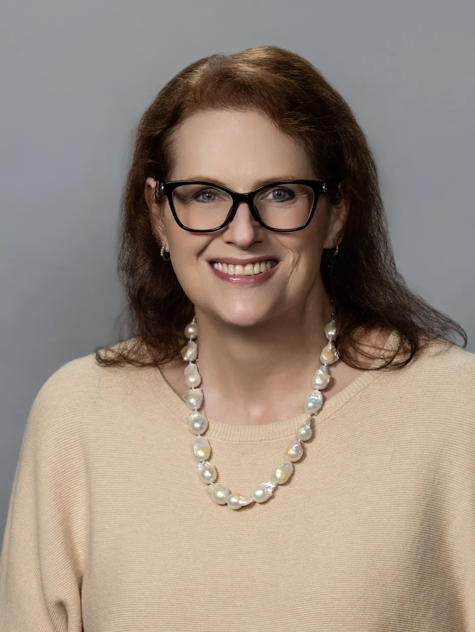 A smiling person with glasses wearing a beige top and a pearl necklace, set against a gray background.