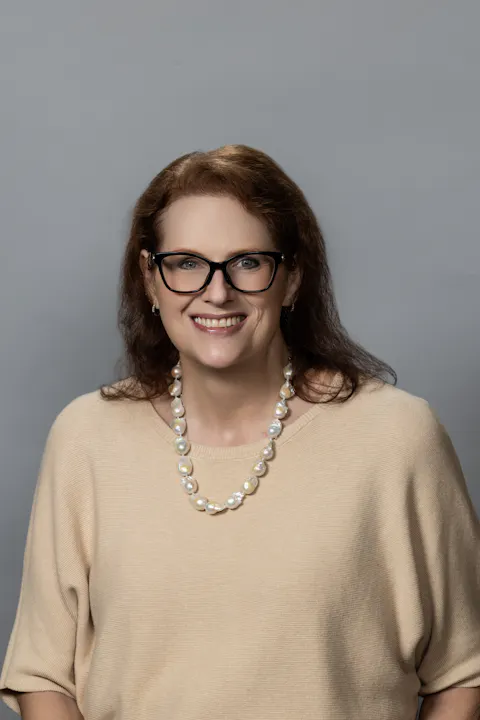 A smiling person with glasses wearing a beige top and a pearl necklace, set against a gray background.