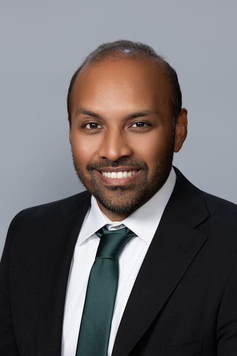The image shows a man with a beard smiling while wearing a suit and green tie, against a plain gray background.