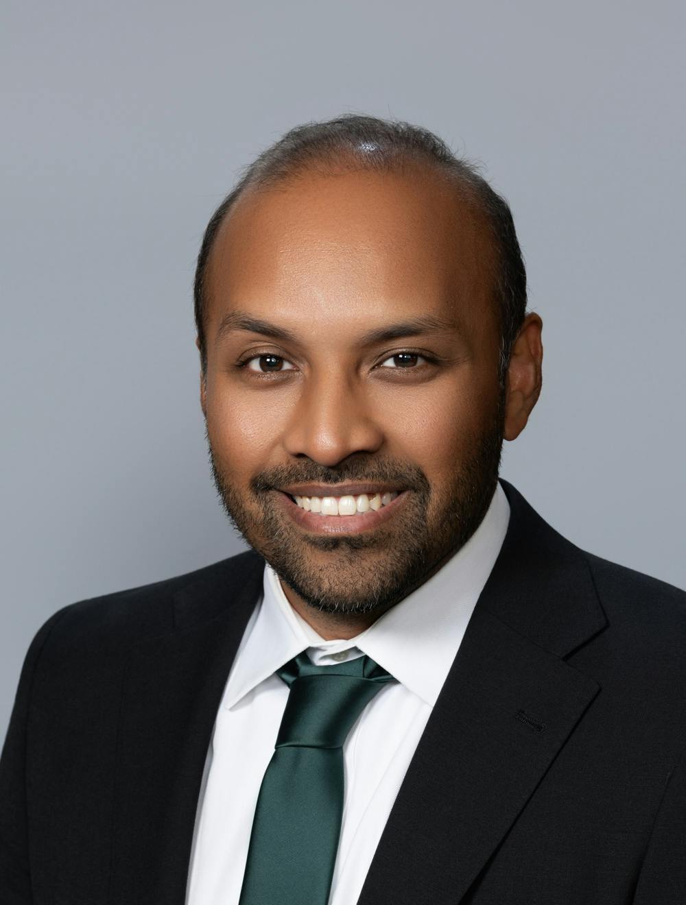 The image shows a man with a beard smiling while wearing a suit and green tie, against a plain gray background.