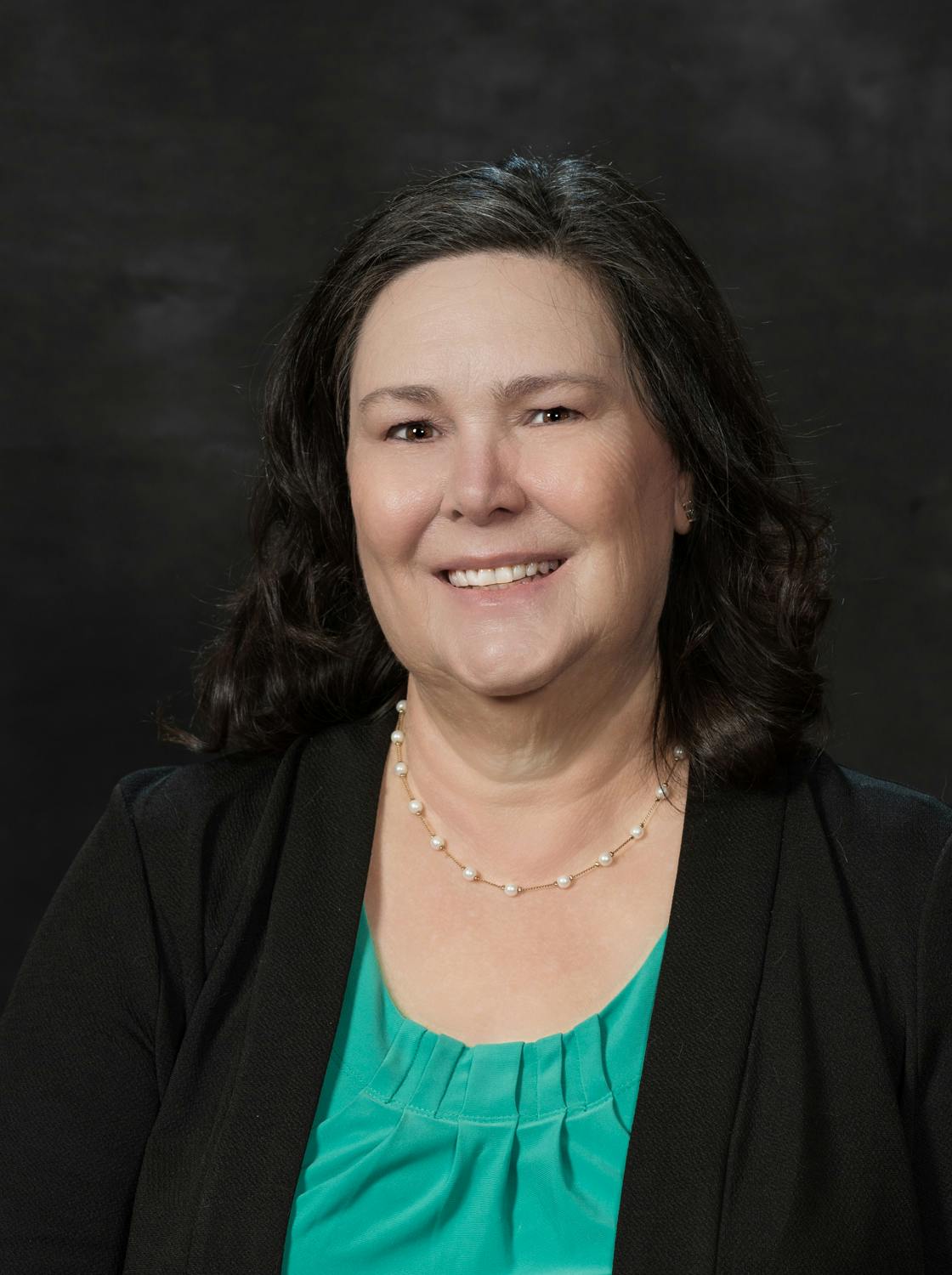 A smiling woman with a necklace, wearing a blazer and a green top, against a dark background.