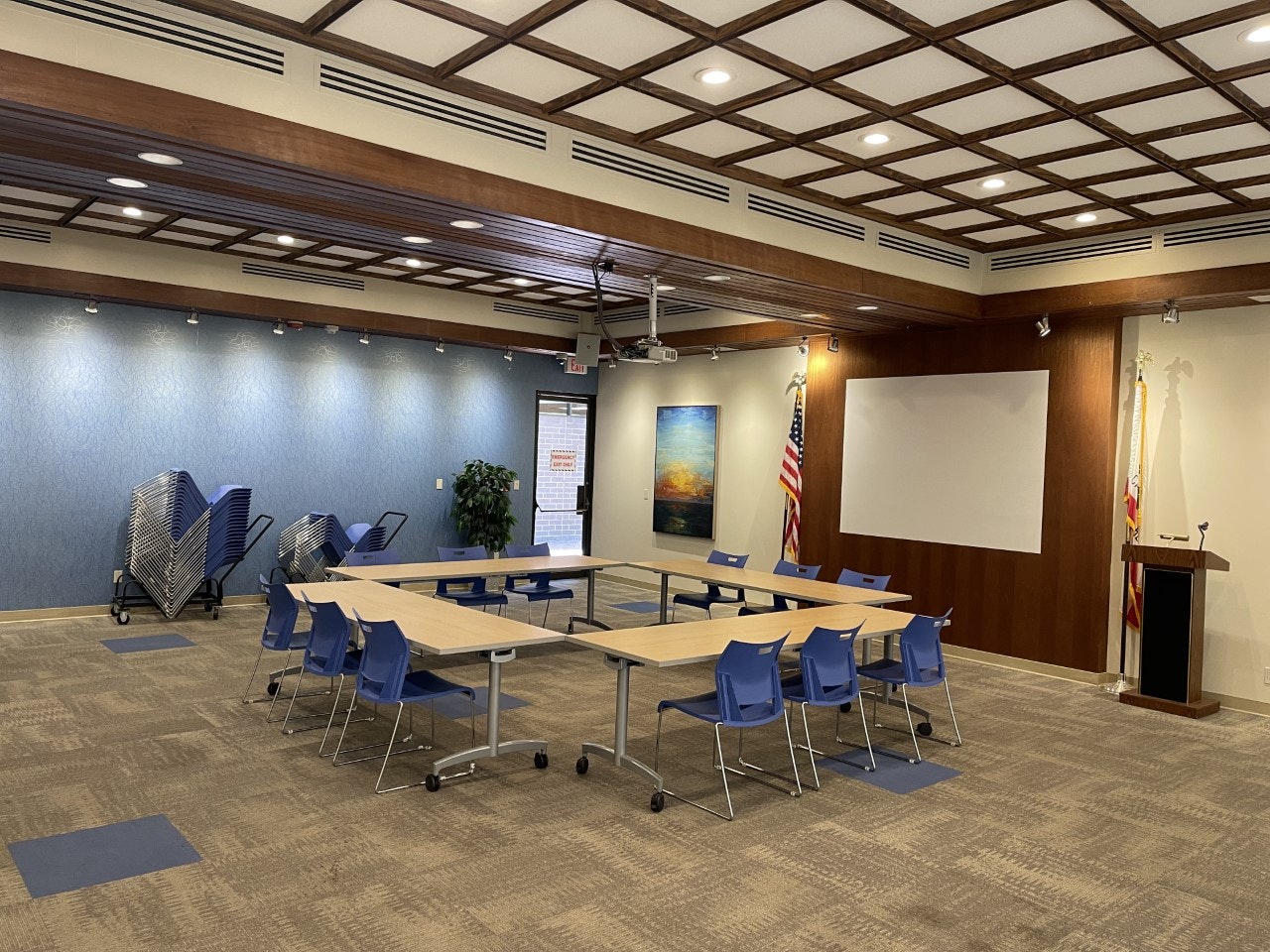 A conference room with square-shaped table setup, blue chairs, projector, American flag, and stacked extra chairs.