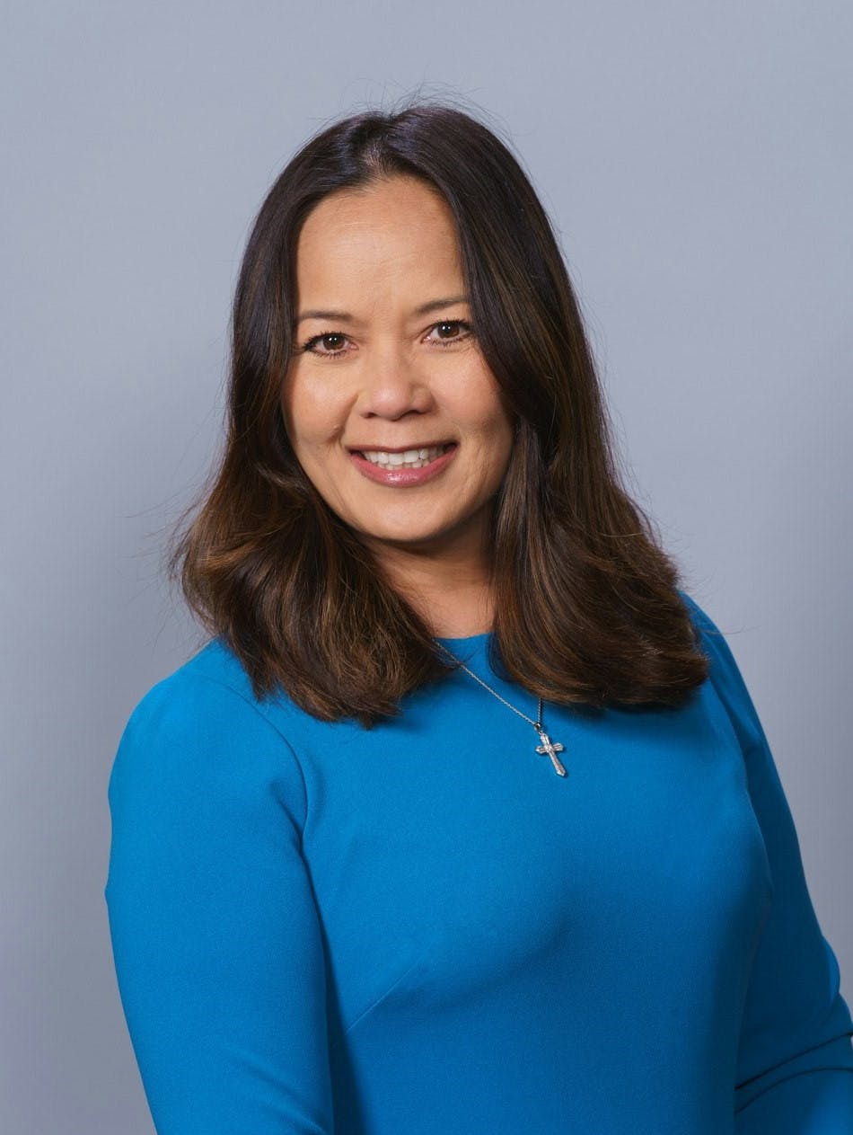 A smiling woman wearing a blue top and a cross necklace against a grey background.