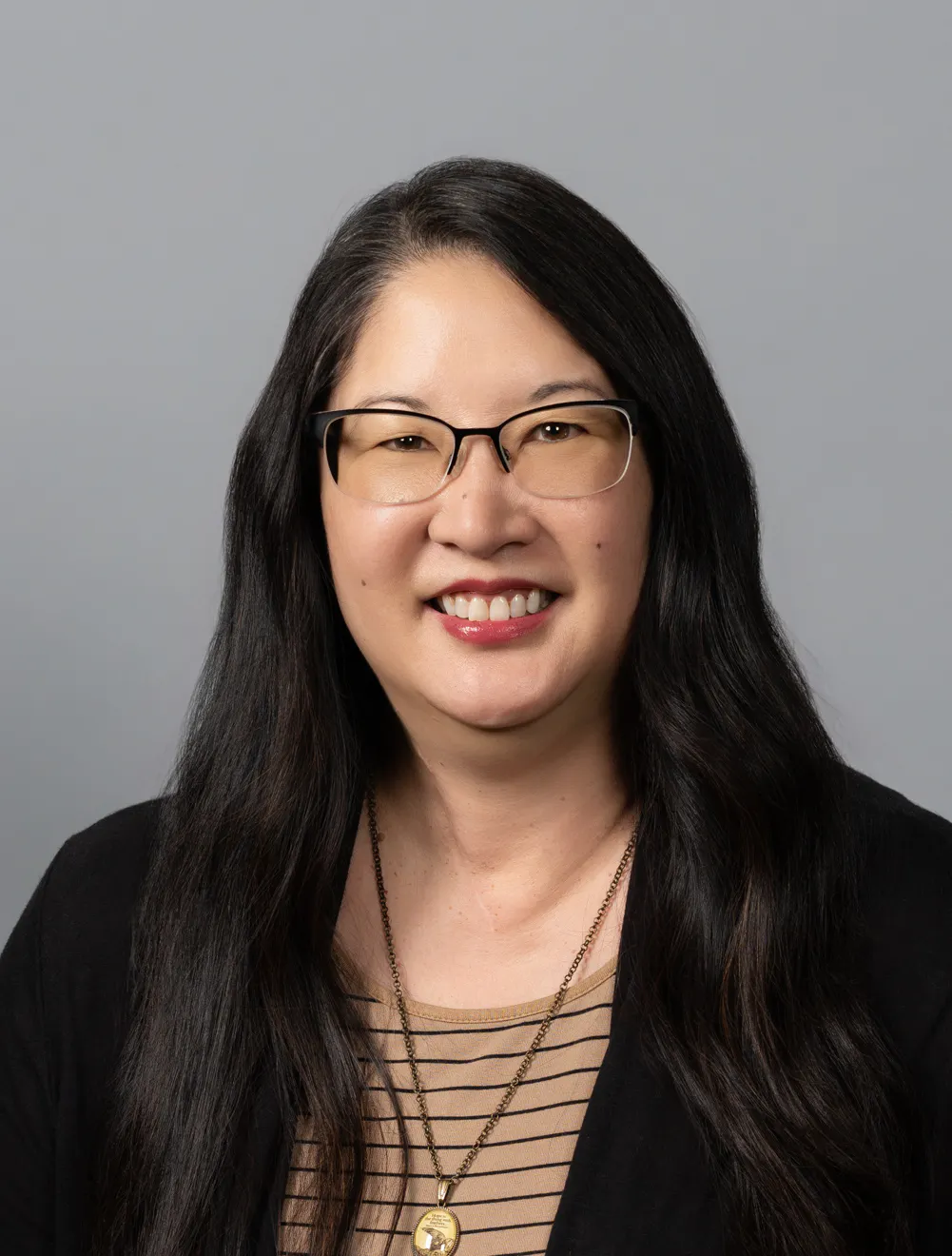 A smiling person with long dark hair, glasses, and a striped top, wearing a pendant necklace.