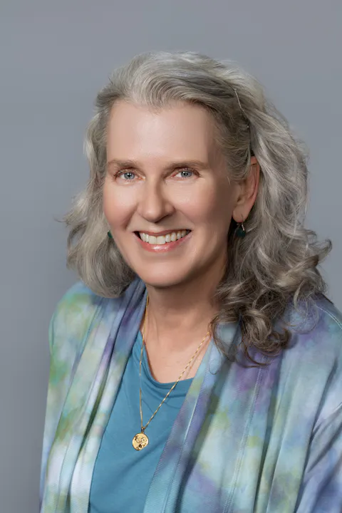 A smiling woman with gray hair poses against a gray background, wearing a colorful shawl and gold jewelry.