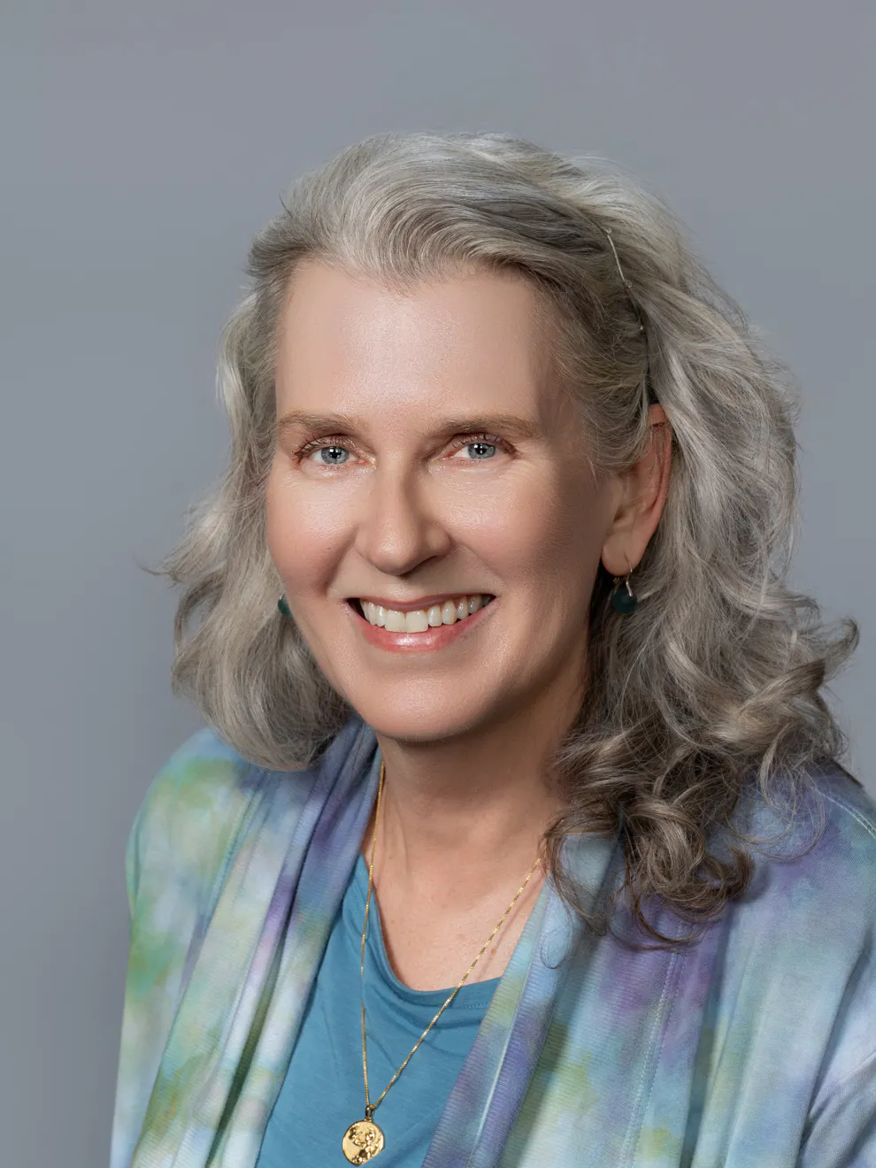 A smiling woman with gray hair poses against a gray background, wearing a colorful shawl and gold jewelry.