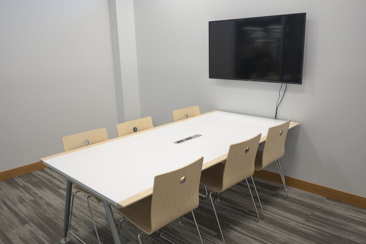 A meeting room with a white table, six wooden chairs, and a wall-mounted TV.