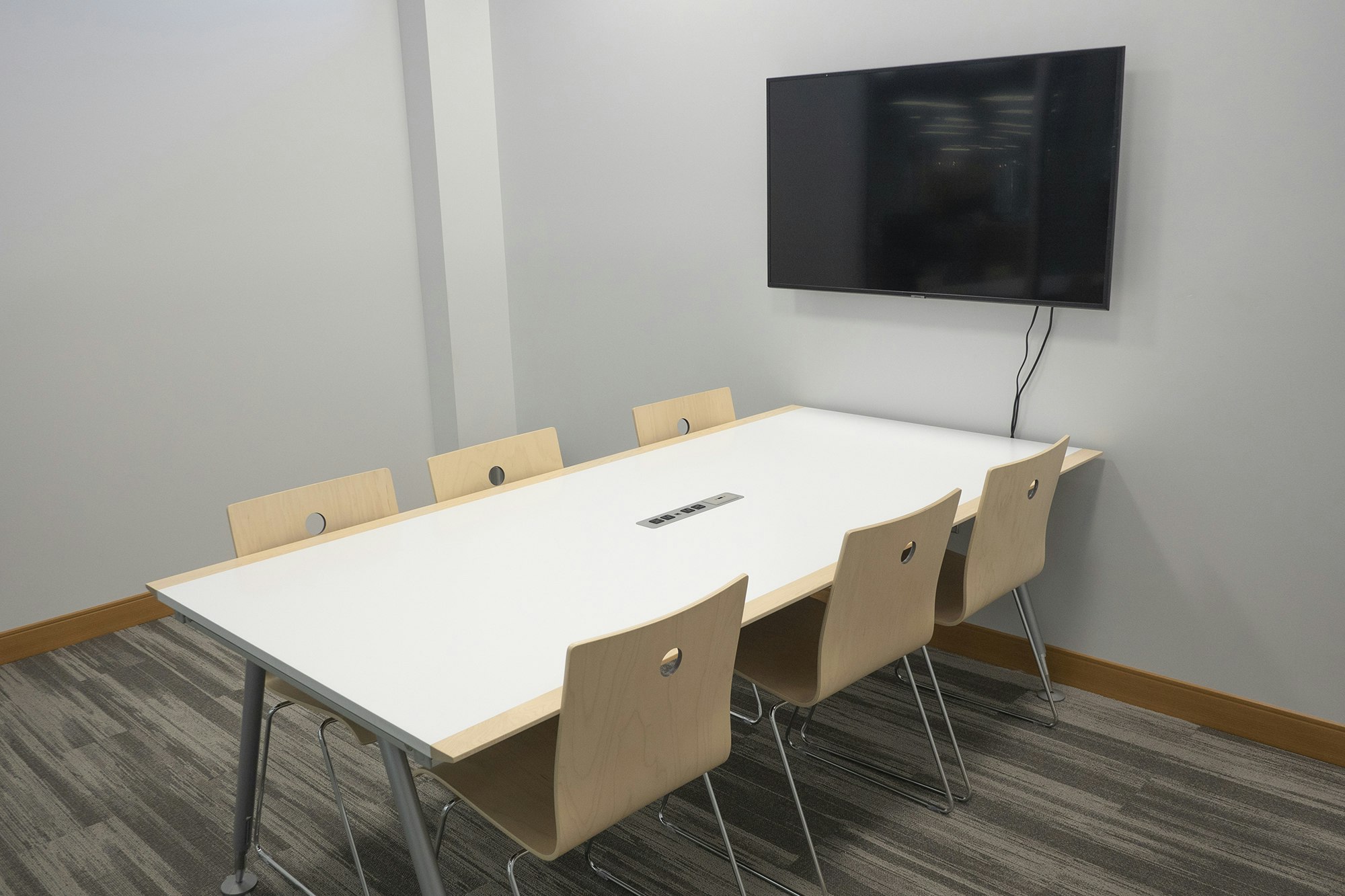 A meeting room with a white table, six wooden chairs, and a wall-mounted TV.