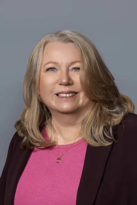 A woman with long blonde hair, wearing a pink top and a dark jacket, smiles against a gray background.