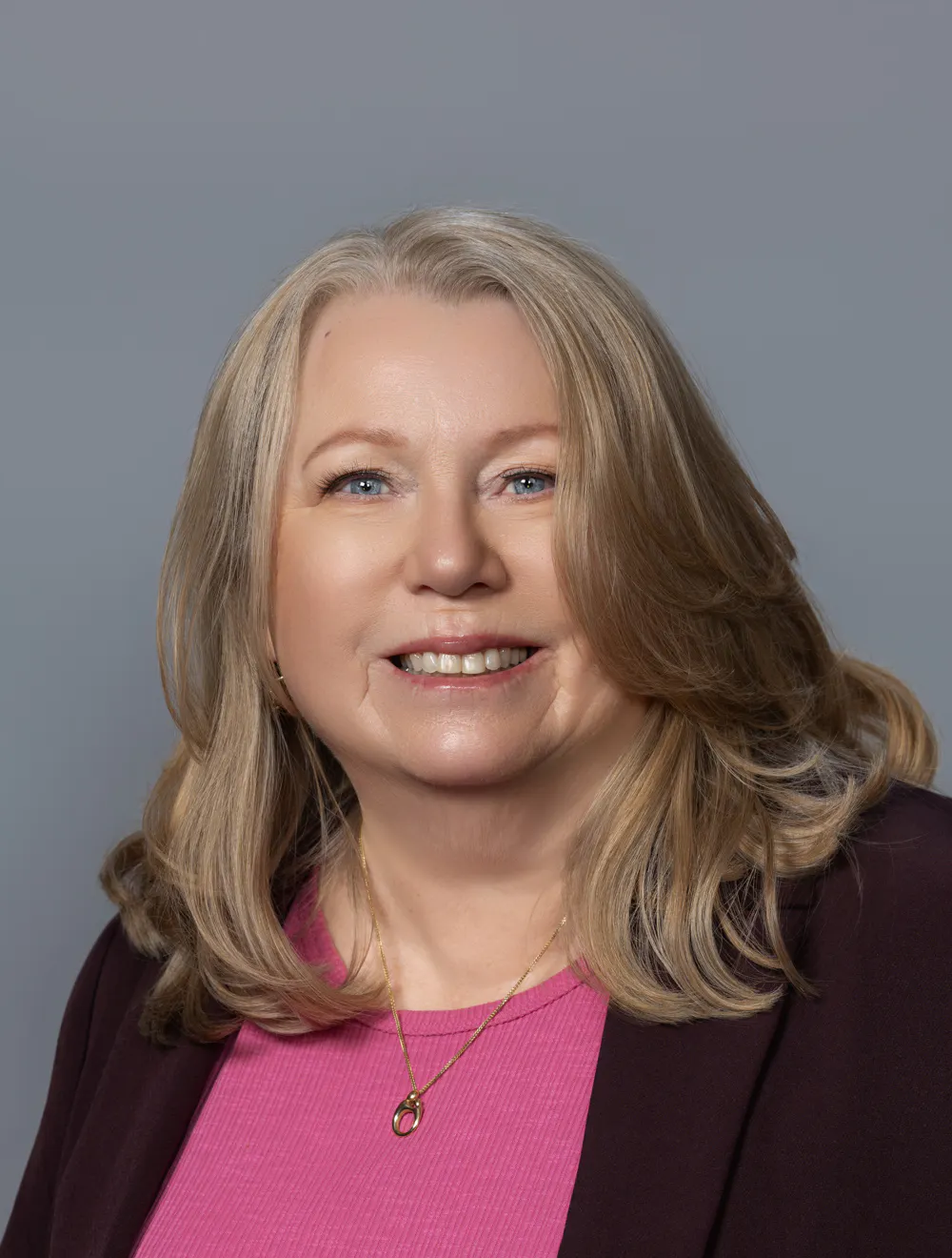 A woman with long blonde hair, wearing a pink top and a dark jacket, smiles against a gray background.