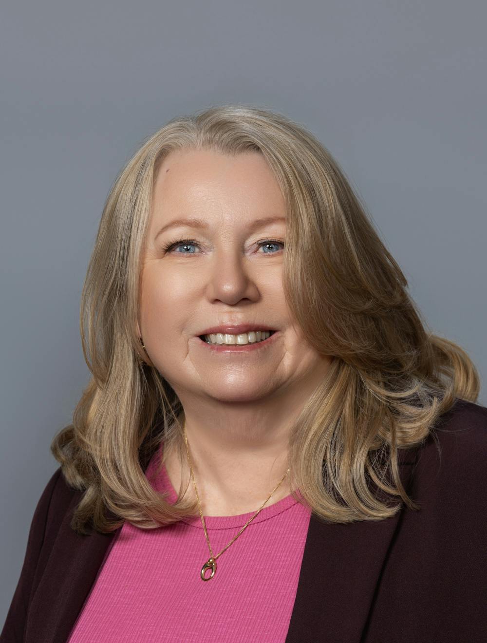 A woman with long blonde hair, wearing a pink top and a dark jacket, smiles against a gray background.