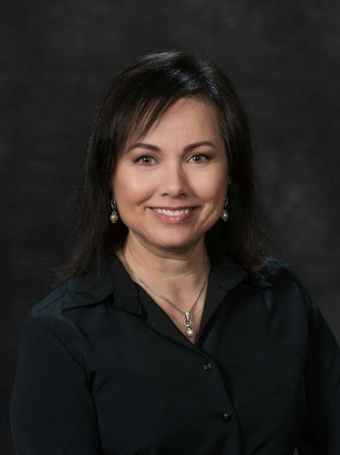A smiling woman with dark hair wearing a black blouse and a pearl necklace against a dark backdrop.