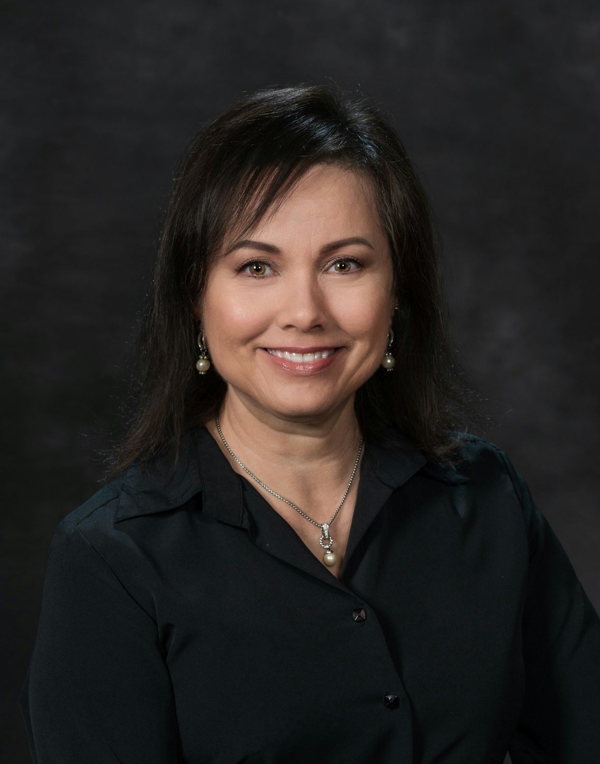 A smiling woman with dark hair wearing a black blouse and a pearl necklace against a dark backdrop.