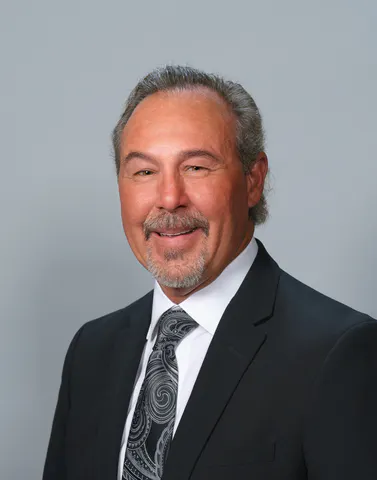 A man with a friendly smile, wearing a black suit and tie against a grey background.