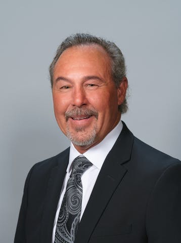A man with a friendly smile, wearing a black suit and tie against a grey background.
