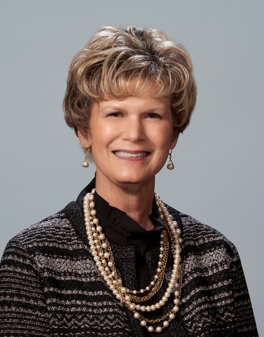 A smiling woman with short hair, wearing a black and white patterned outfit, adorned with layered pearl necklaces.