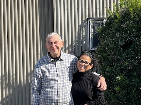 Two people smiling, embracing outside by a corrugated wall and greenery under sunny skies.