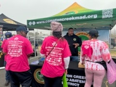 Group of people in pink shirts at a booth with a banner reading "www.cottoncreekmad.org". Event or community gathering.