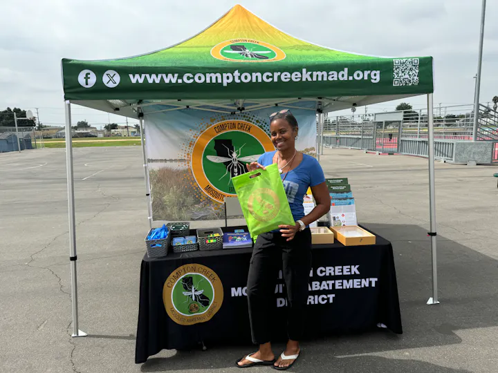 A smiling person stands by an info booth with a banner, branded materials, and a green tote bag, likely at an outdoor event.