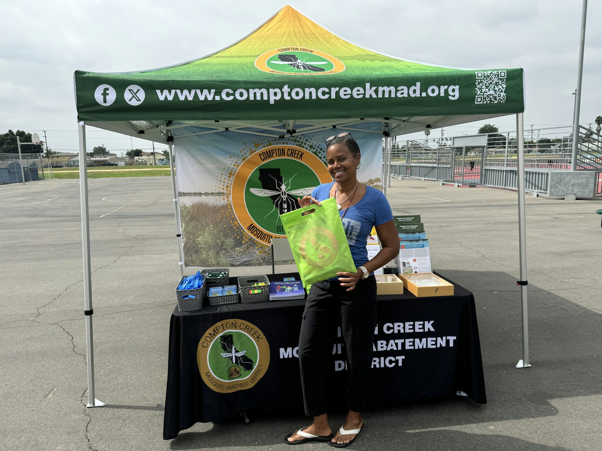 A smiling person stands by an info booth with a banner, branded materials, and a green tote bag, likely at an outdoor event.